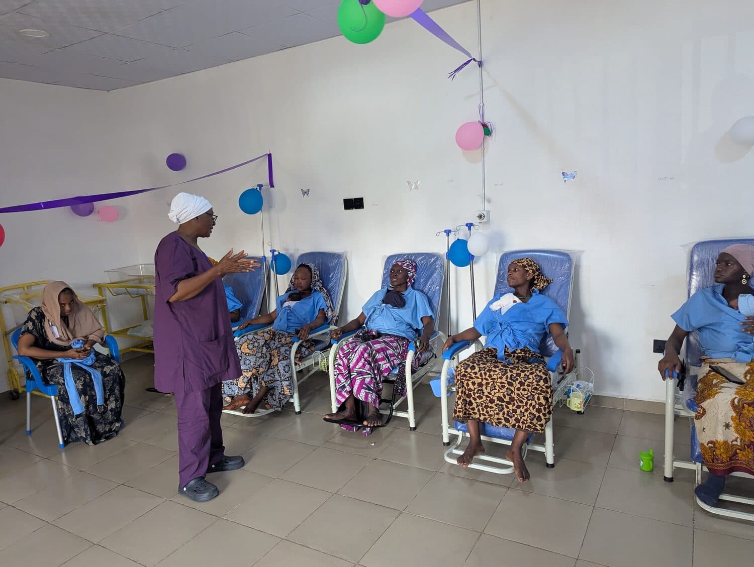 A nurse counselling a row of mothers in Kangaroo Care chairs, each holding her newborn