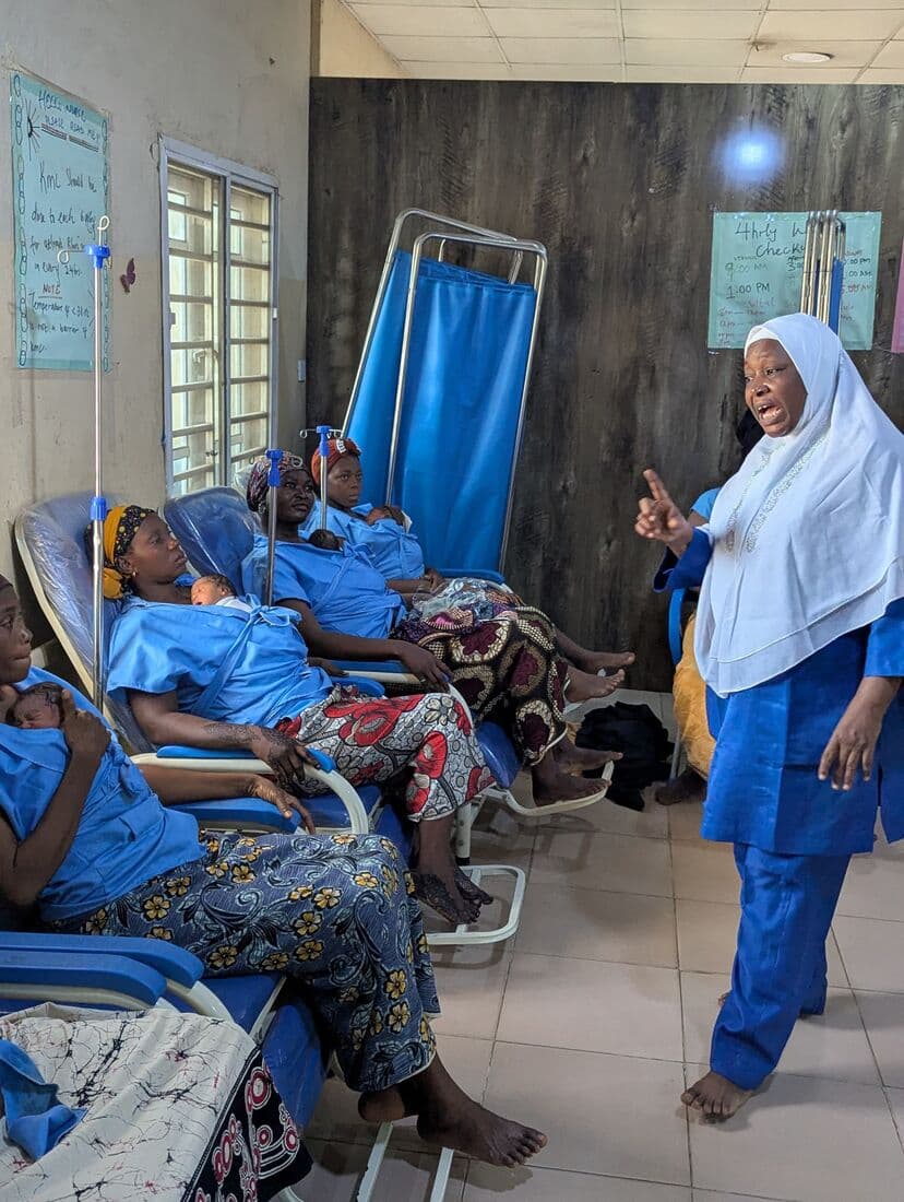 A nurse talking the ward through Kangaroo Care, mothers holding their newborns in the chairs