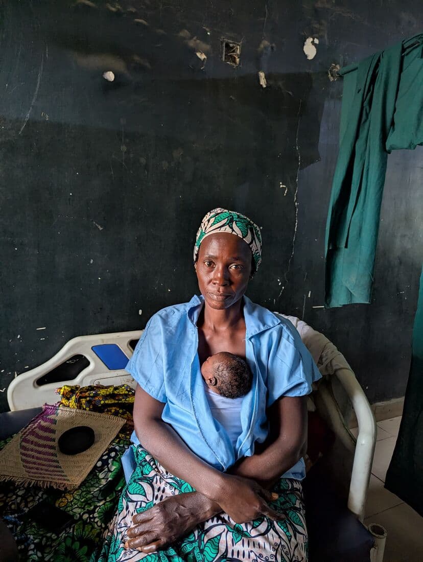 A mother seated at the end of the ward with her newborn held against her chest