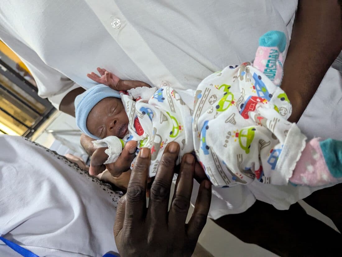 A newborn in a patterned wrap held in a nurse's hands on the ward